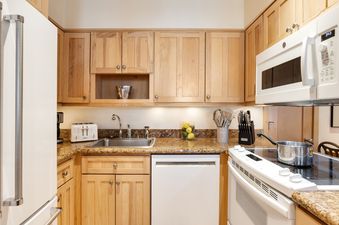 A modern kitchen with light wood cabinets, granite countertops, and white appliances, including a refrigerator, sink, dishwasher, oven, and microwave, at Tamarack 01.