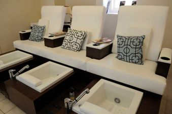 A row of three comfortable white chairs, each with a pillow and a small table featuring a cup holder, faces individual white foot basins set into dark wood bases, creating a serene and inviting spa-like atmosphere.
