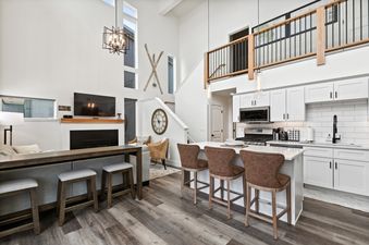 A modern open-concept living area with a white kitchen, an island with bar stools, a living room with a fireplace and TV, and a loft overlooking the space.