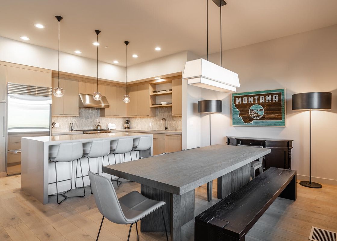 The dining area of Moonlight Basin 80B Lodgeside features a large, grey table with a black bench and chairs, a view of the kitchen, and a wall with a rustic Montana sign.