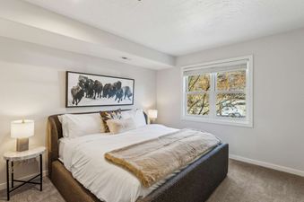 A cozy bedroom featuring a large bed with white bedding and a faux fur throw, flanked by two nightstands with lamps, and a window offering a view of outdoor trees.