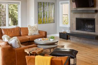 A cozy, rustic-modern living room featuring a brown leather sectional, unique coffee tables, birch tree wall art, a fireplace, and a large window with an outdoor view.