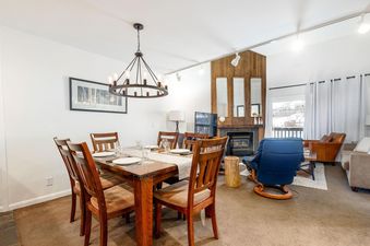 A wide view of the dining area and an adjacent living room at Snow Flower 129 Park City, featuring a wooden dining table and chairs, a fireplace, and comfortable seating.