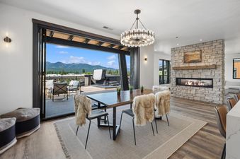 The dining area at Grandview Escape, with a wooden table, faux-fur adorned chairs, and a large open-air view of mountains and a deck with a hot tub.
