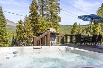 An expansive outdoor deck at Grand View on the Blue features a hot tub in the foreground, with a dining area, BBQ grill, and stunning mountain and forest views in the background.