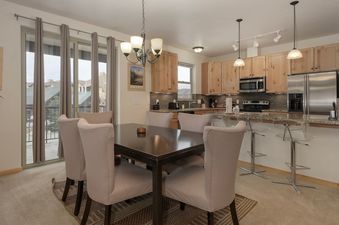 The dining area and kitchen of Trailhead Lodges 422 are elegantly furnished, featuring a large dark wood dining table surrounded by six comfortable light-colored chairs, and a modern kitchen with light wood cabinetry, stainless steel appliances, and a gra