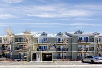 A modern multi-story apartment building with olive-green siding, black balconies, and a light-colored roof under a blue sky with wispy clouds.