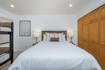 A cozy bedroom at River Street Refugio featuring a queen bed with white linens, decorative pillows, bedside lamps, a wooden headboard, and a black bunk bed to the side.