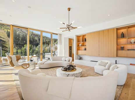 A bright and airy living room at Founders Place 1704, featuring curved cream sofas, marble coffee tables, modern lighting, and large glass doors that open to a balcony with mountain views.