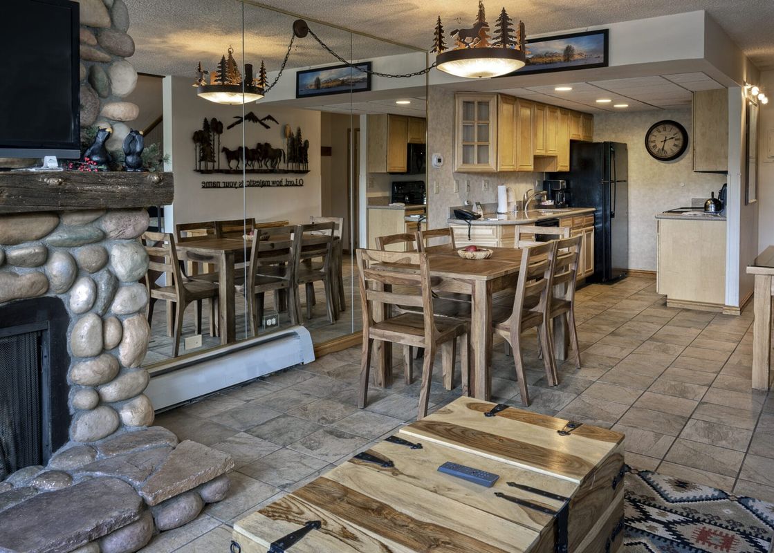 A spacious living area in the Two Bedroom Suite Beaver Run Resort, featuring a stone fireplace, a flat-screen TV, a rustic wooden dining table, and a well-equipped kitchen with light wooden cabinets and modern appliances.