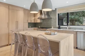 A modern kitchen with light wood cabinetry, a large kitchen island with clear bar stools, and stainless steel appliances.