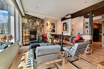 A modern and artistic living room featuring a brown leather sectional, grey armchairs, a stone fireplace, large windows with a view, and vibrant abstract art.