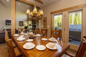 The dining area at Leland Creek Home features a large wooden table set with white dishes, surrounded by wooden chairs, under a rustic chandelier, with a view of a hot tub through glass doors.