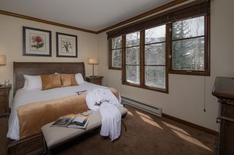 The bedroom in Chalet Fortessa features a large bed, two bedside tables with lamps, and a window offering a view of the snow-covered trees and distant mountains.