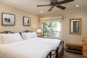 A cozy bedroom in Montaneros 406 features two beds with wrought iron frames, white bedding, and framed bear artwork, with a window offering a view of green trees.