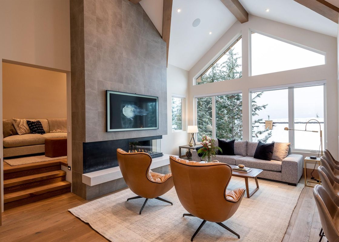 A luxurious living room in Northern Lights 32 featuring a large fireplace with a TV mounted above, two brown leather armchairs, a grey sofa, and expansive windows offering a snow-covered mountain view.
