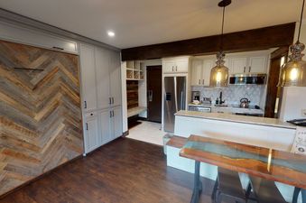 A kitchen with stainless steel appliances, white cabinets, and a unique wooden table with a clear epoxy resin top.  The kitchen is open to a living area with a grey cabinet that slides to reveal a herringbone patterned wall.