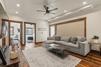 A spacious and inviting living room at Baker Loft 201 features an L-shaped gray sectional sofa, a modern black coffee table, and a patterned area rug, with wooden floors and recessed lighting.