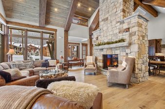 A spacious and luxurious living room with a large stone fireplace, comfortable sofas, and big windows offering a view of a snowy winter landscape.