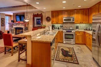 A modern kitchen with stainless steel appliances and wooden cabinets, a stone fireplace in the background, and a dining area with a wooden table and chairs.