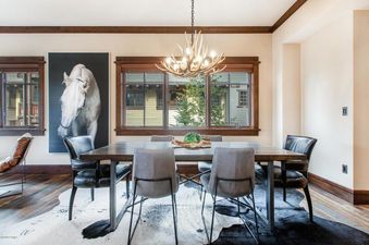 A rustic yet refined dining area at Arrowleaf Lodge 109, featuring a dark wood table with a mix of leather and gray fabric chairs, under an antler chandelier, with a cowhide rug and a large window overlooking green trees.