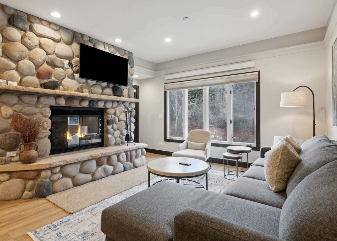 A cozy living room at The Charter D3105 with a large gray sectional sofa, a stone fireplace with a TV, a round coffee table, and windows overlooking a snowy winter landscape.