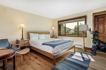 A bedroom with a queen-sized bed, a wooden nightstand with a lamp, a chair, a small table, and a window with a view of a forest. There is an exercise bike in the corner of the room.