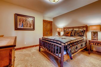A rustic bedroom at Bear Tracks Lodge features a log bed with patterned bedding, two nightstands with lamps, a wooden dresser, and framed artwork on the wall.