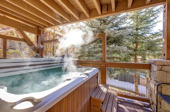 A steamy hot tub on a wooden deck with a scenic view of snow-dusted trees and fall foliage, framed by wooden beams and a lattice railing.