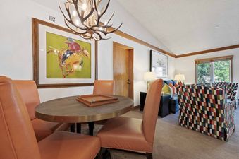 A cozy dining area featuring an oval table with orange leather chairs under an antler chandelier, transitioning into a living area with colorful seating and a window overlooking nature in The Charter A4505.