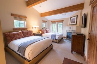A spacious bedroom within The Chateaux Four Bedroom Suite, featuring two large beds with elegant bedding, a stone fireplace, and multiple windows offering views of the outdoors.