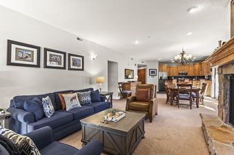 A wide view of the comfortable and inviting living room at Town Pointe C101, showing a blue sofa, a rustic coffee table, and an adjacent dining area and kitchen.