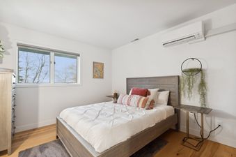 The master bedroom at Stumptown House features a large bed with a wooden headboard, white bedding, and decorative pillows, a large window, a modern side table, and an air conditioning unit.