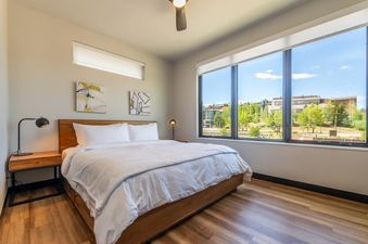 The primary bedroom at Luxury Alpine Abode features a comfortable bed with white linens, two bedside tables with lamps, and large windows overlooking the scenic landscape.