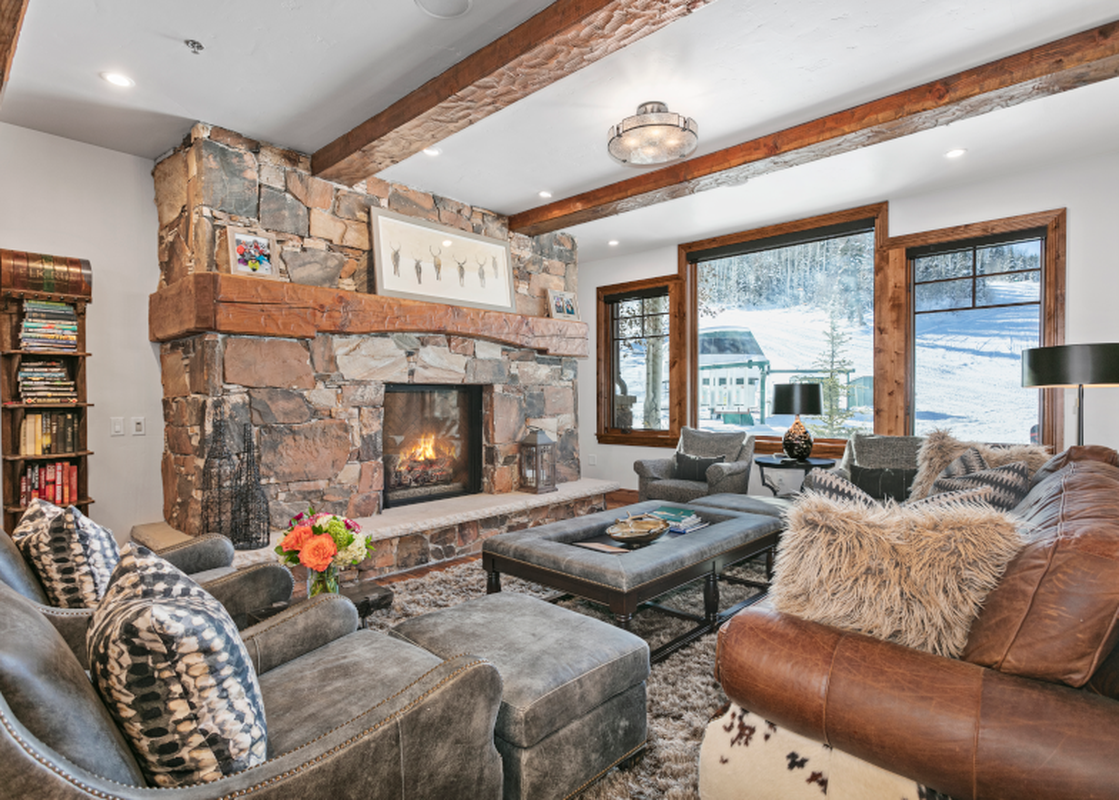 The spacious living room of Grand Lodge 3 Bedroom Deluxe 304 features a cozy fireplace, comfortable leather furniture, and a breathtaking view of the snowy mountain slopes outside. The room has a contemporary design and is well-lit by natural sunlight. Th