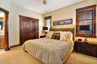 A king-sized bed with a patterned duvet, throw pillows, and two dark wooden nightstands with lamps, framed by windows with blinds and wooden doors, creating a cozy bedroom space.