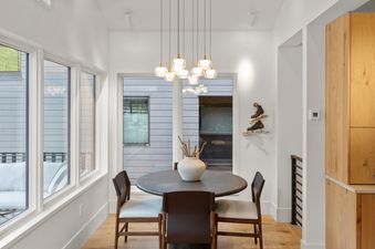 A modern dining area features a dark circular table with four chairs, illuminated by a cluster of pendant lights, with large windows overlooking an outdoor view.