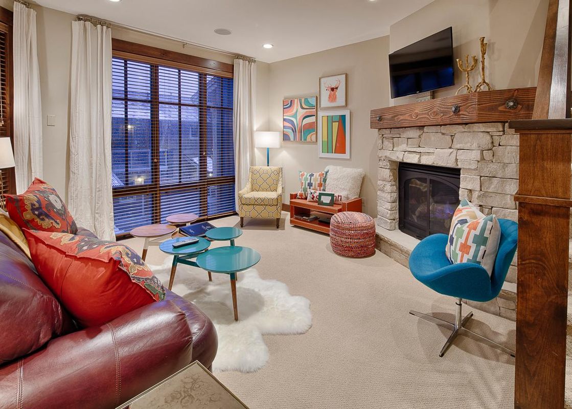 The vibrant living area of Silver Star 702, featuring a red leather sofa, colorful accent chairs, a stone fireplace, and large windows with wooden blinds.