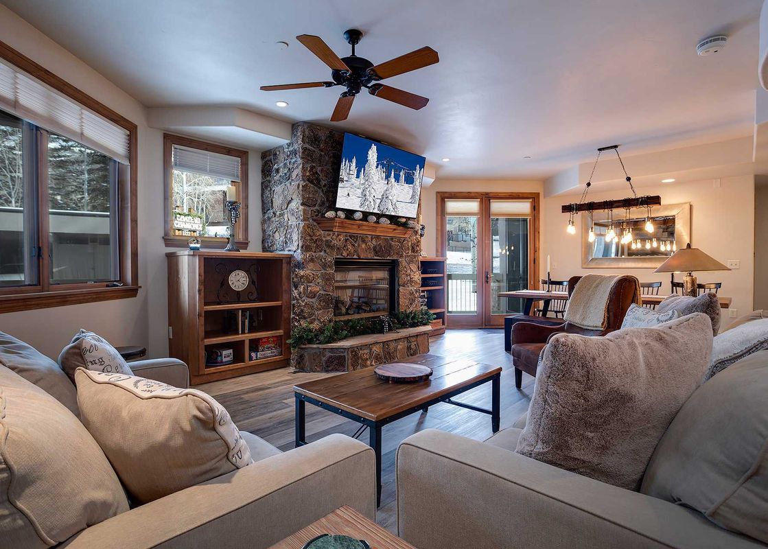 The living room features a stone fireplace, flat-screen TV, comfortable seating, and large windows with views of the outdoors, offering a cozy and inviting space.