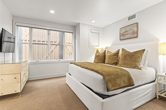 A serene bedroom at Rio Grande 203 features a large bed with white linens and gold accents, a flat-screen TV above a modern dresser, and a window overlooking a wooden fence.