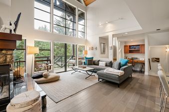 A bright and spacious living room at Whistler Pinnacle Ridge 42 features an L-shaped gray sectional sofa, a white marble coffee table, and large windows offering views of green trees and a balcony.
