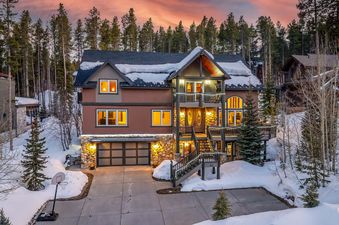 The Ski Hill Sanctuary is a beautiful home set against a snow-covered landscape and a forested backdrop under a sunset-colored sky.