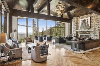 Spacious great room at Grandview at The Colony with high wood-beamed ceilings, a large stone fireplace, comfortable seating, and massive windows offering mountain views.