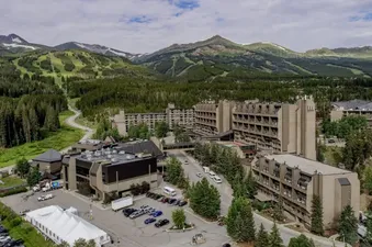 An aerial view of The Summit Suite Beaver Run Resort with several large buildings, a parking lot with a white tent, and a road winding through a dense forest with mountains and ski slopes in the background under a cloudy sky.