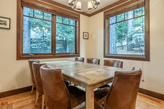 A rustic dining room at Cedar Crest Owl Cottage 9 with a large wooden table and six brown leather chairs, framed by windows looking out onto a forest.