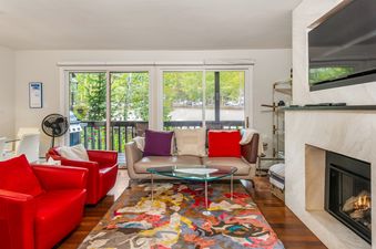 A spacious living room features two red armchairs, a gray sofa with colorful throw pillows, and a glass coffee table, all set on a vibrant patterned rug, with a large fireplace and television to the right and sliding glass doors leading to a balcony on th