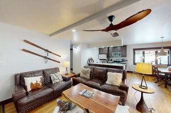 The living room features a large leather couch with throw pillows, a coffee table with a magazine on top, and a ceiling fan. There are two additional sofas and a side table with a lamp.