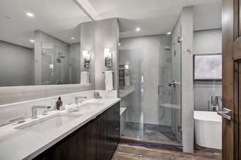A modern bathroom at Snow Cloud Lodge 506 with a double vanity, a glass-enclosed shower, and a freestanding bathtub, featuring sleek gray tiles and rich wood flooring.