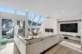 A modern and bright living room at Luxe Lookout featuring a large sectional sofa, a sleek fireplace, a flat-screen TV, and expansive windows with views of snow-covered mountains.
