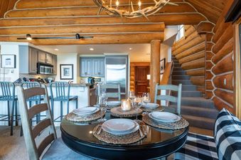 A dining table with four place settings set with plates, cutlery, and glasses, is in the center of the image.  There is a large rustic wooden chandelier above the table.  The table is situated in a log cabin with a stairway leading to a loft. The kitchen